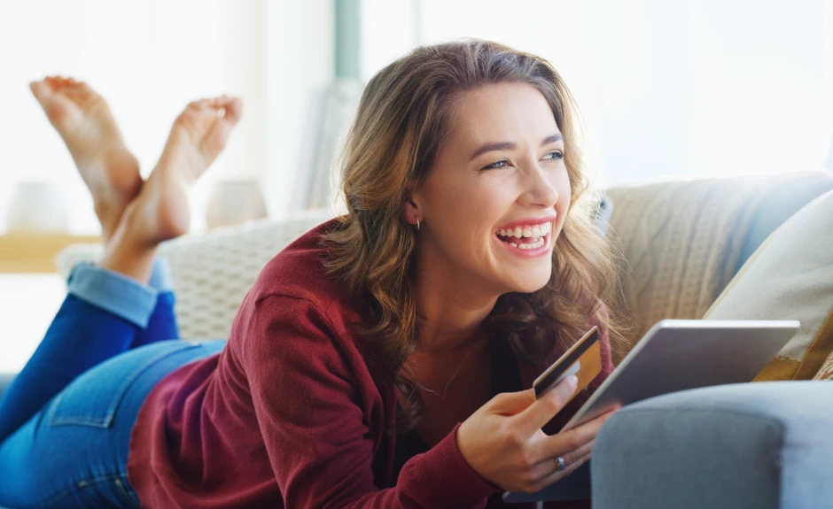 Frau liegt lachend auf der Couch und hat eine Kreditkarte, sowie ein Tablet in der Hand.