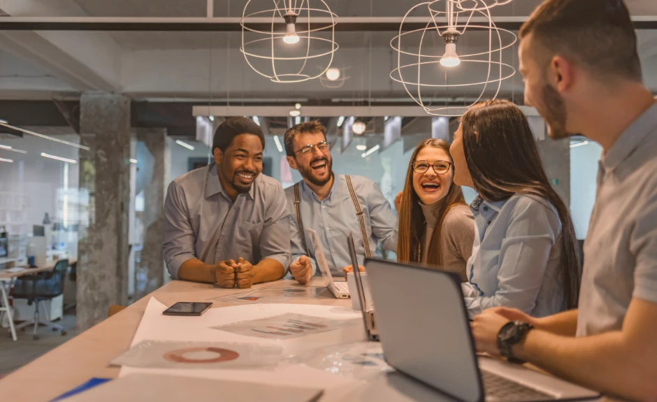 Fünf Personen stehen um einen Tisch im Büro und lachen. Auf dem Tisch liegen Dokumente und Laptops.