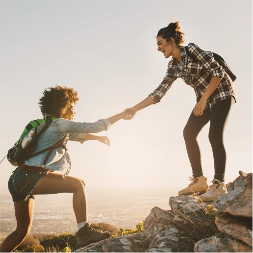 Freunde werben Freunde TF Bank Aktion: 2 Freundinnen helfen sich, um auf einen Berg zu gelangen. Eine Frau hält der anderen den den Arm zur Hilfe hin. Im Hintergrund geht die Sonne auf.