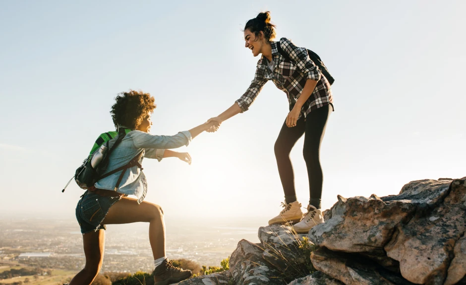 Freunde werben Freunde Aktion: 2 Freundinnen helfen sich, um auf einen Berg zu gelangen. Eine Frau hält der anderen den den Arm zur Hilfe hin. Im Hintergrund geht die Sonne auf.