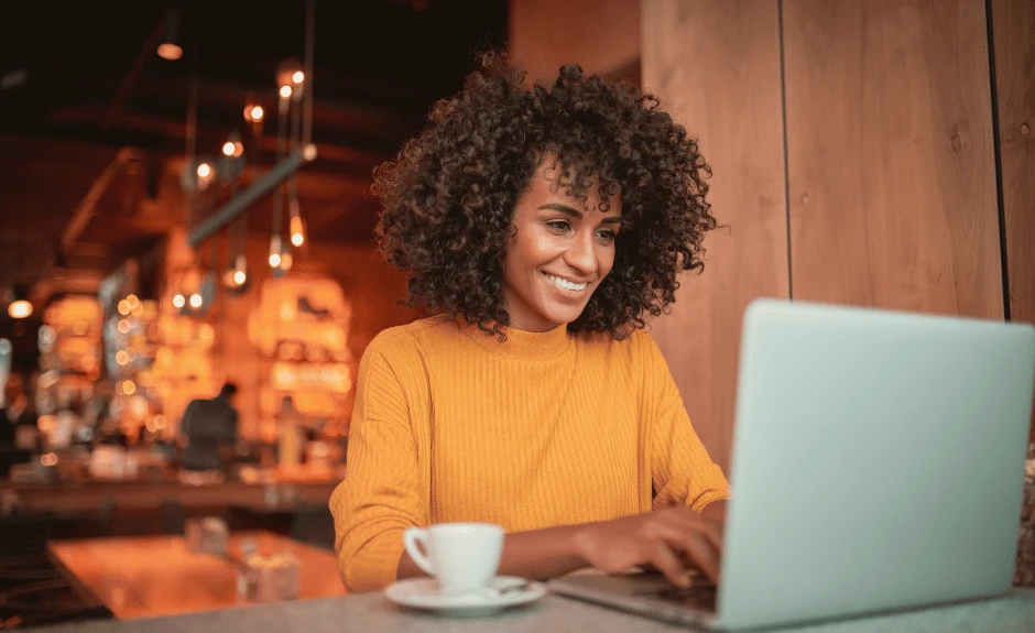 Frau sitzt am Tisch und tippt lächelnd etwas in ihren Laptop. Danben steht ein Kaffee.